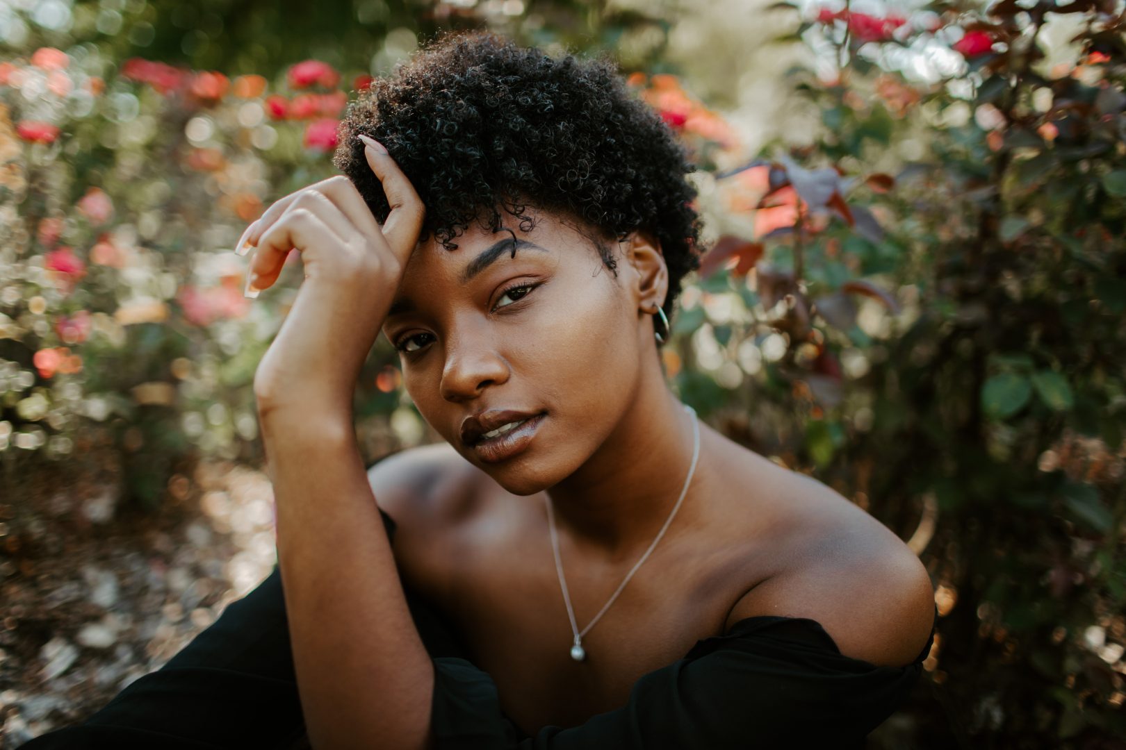 A woman wearing a black off-the-shoulder dress setting among out-of-focus rose bushes.