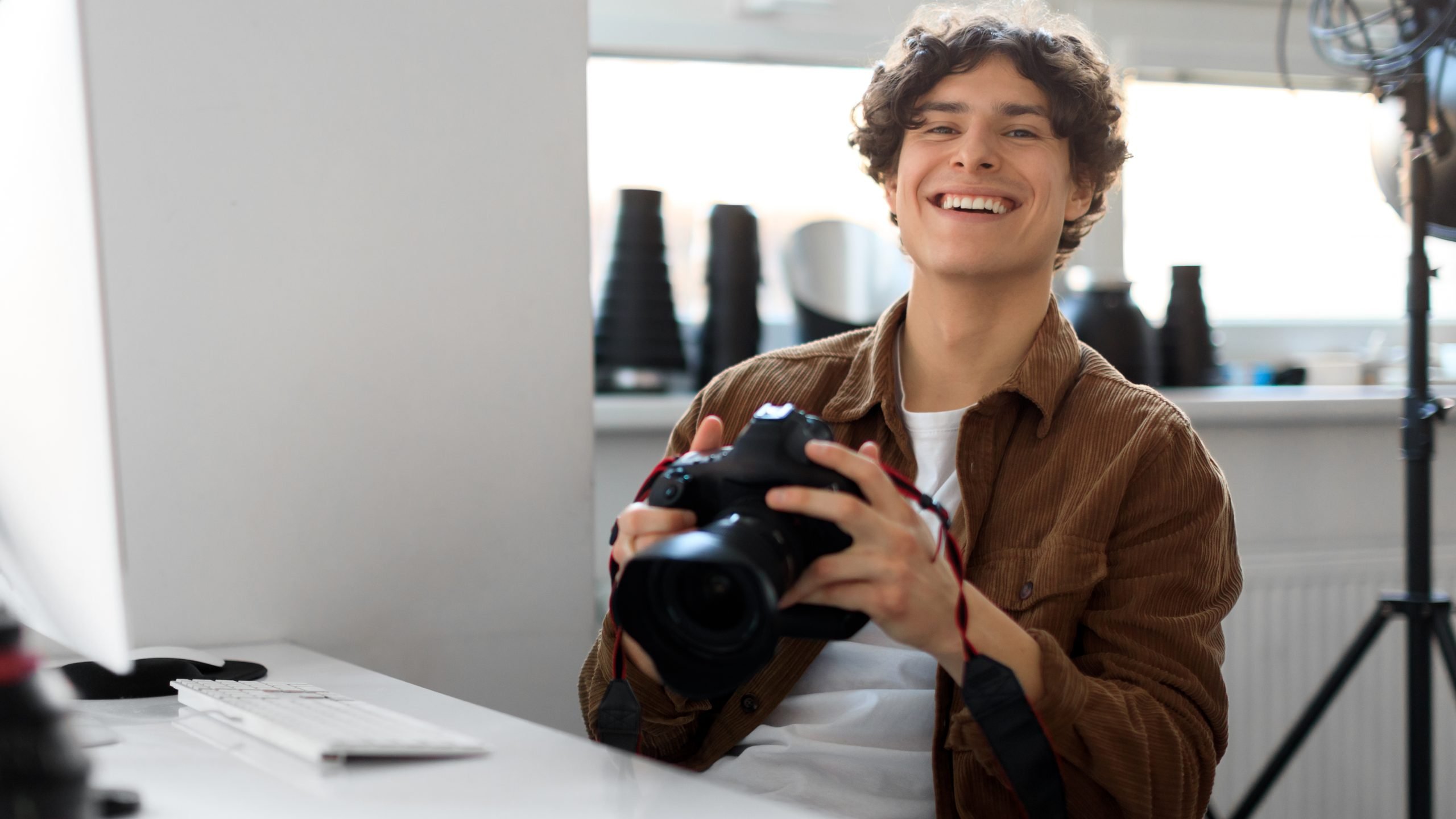 Professional photographer young man working on photo shoot at studio, holding dslr camera, using pc computer, looking and smiling at camera, copy space